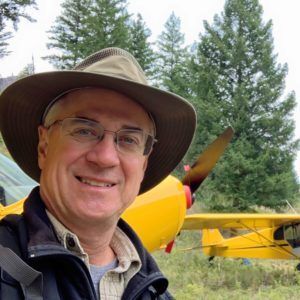 A man wearing a hat and glasses is standing in front of a yellow plane.
