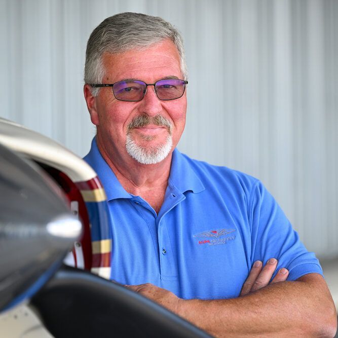 Man with gray hair and glasses in a blue shirt, arms crossed, standing near a propeller plane in a hangar.