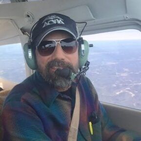 A man with a beard is sitting in a plane wearing headphones and sunglasses.