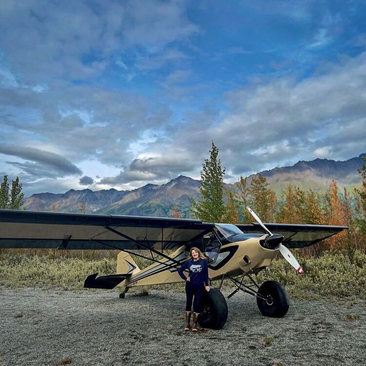 A person standing next to a small plane with mountains in the background