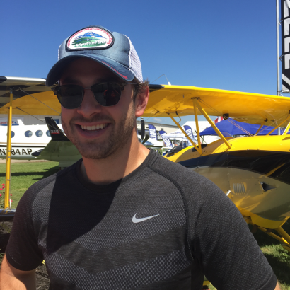 A man wearing a hat and sunglasses is smiling in front of a plane with the letters baap on it