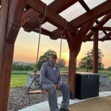 A man is sitting on a wooden swing under a pergola at sunset.