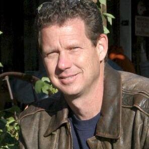 A man wearing a brown leather jacket and a blue shirt is smiling for the camera.