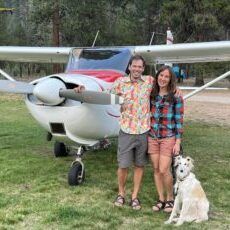 A man and a woman are standing in front of a small plane with a dog.