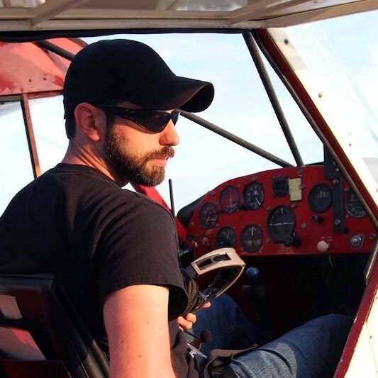 A man wearing sunglasses and a hat is sitting in the cockpit of an airplane