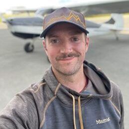 A man wearing a hat and a hoodie is taking a selfie in front of an airplane.