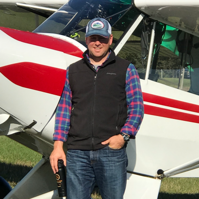 A man in a plaid shirt and black vest is standing in front of an airplane