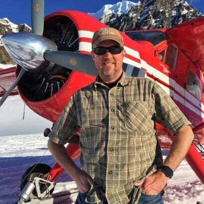 A man in a plaid shirt is standing in front of a red plane.