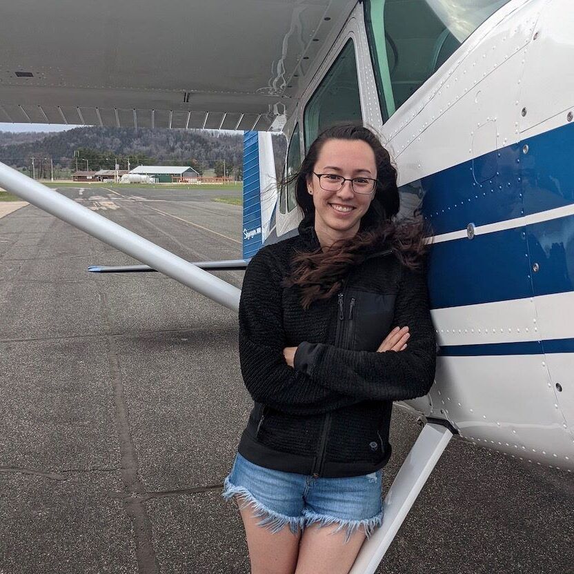 Woman with glasses and arms crossed, stands beside small airplane on a runway.