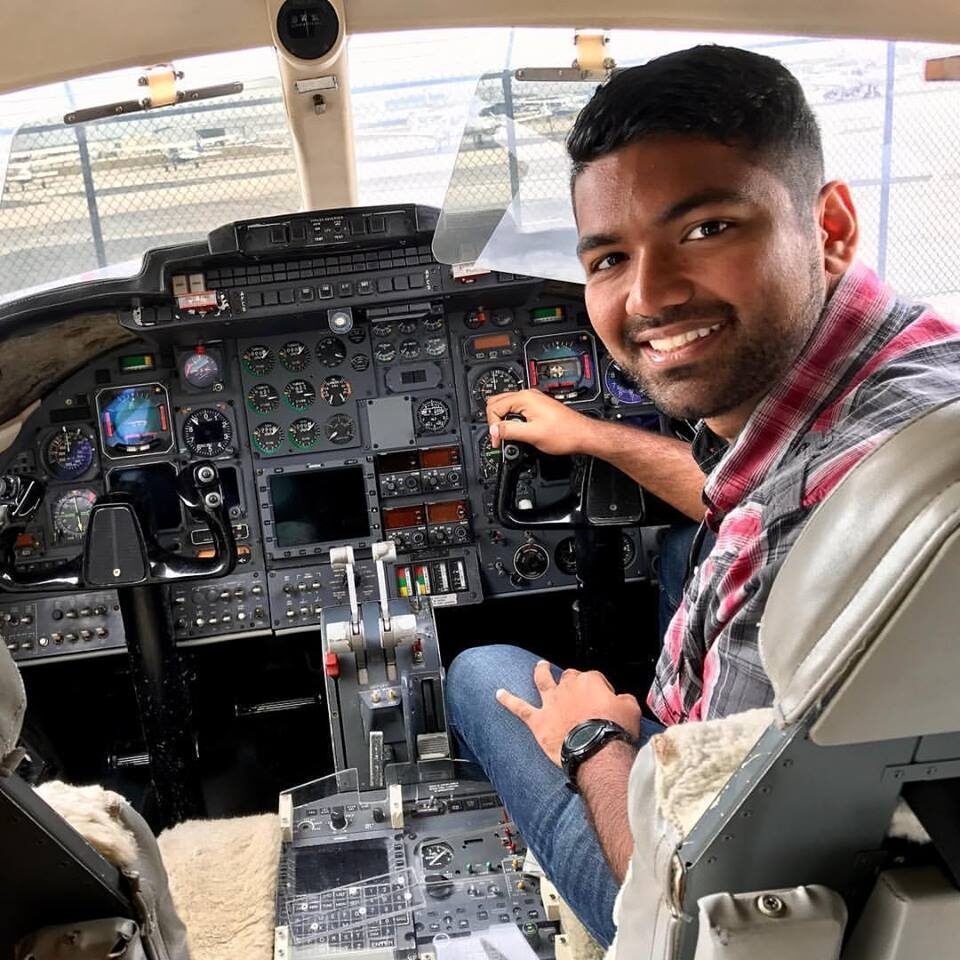 Man in cockpit of plane smiles, adjusting controls; wearing plaid shirt and jeans.