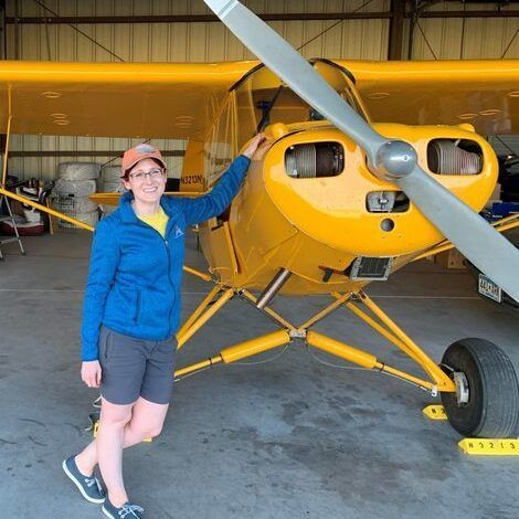 Woman in a blue jacket and hat next to a yellow airplane in a hangar.