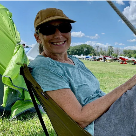 Man in front of brown and red airplane. Smiling with hands on hips. Airport setting.