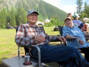 Two men are sitting in chairs in a field with mountains in the background.