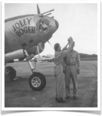 Two men stand in front of a jolly roger airplane