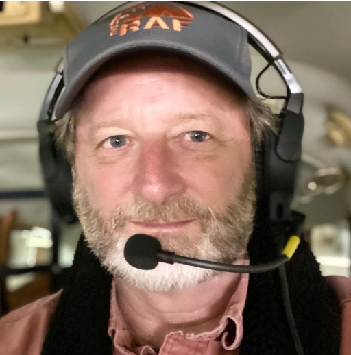 Man in hat and sunglasses in front of a small white airplane on a white plane. Blue sky.