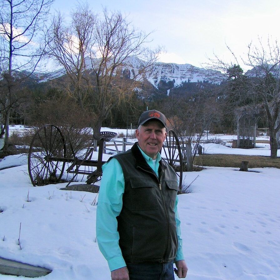 Man in vest and cap, snow-covered yard, antique wheels, mountain backdrop.