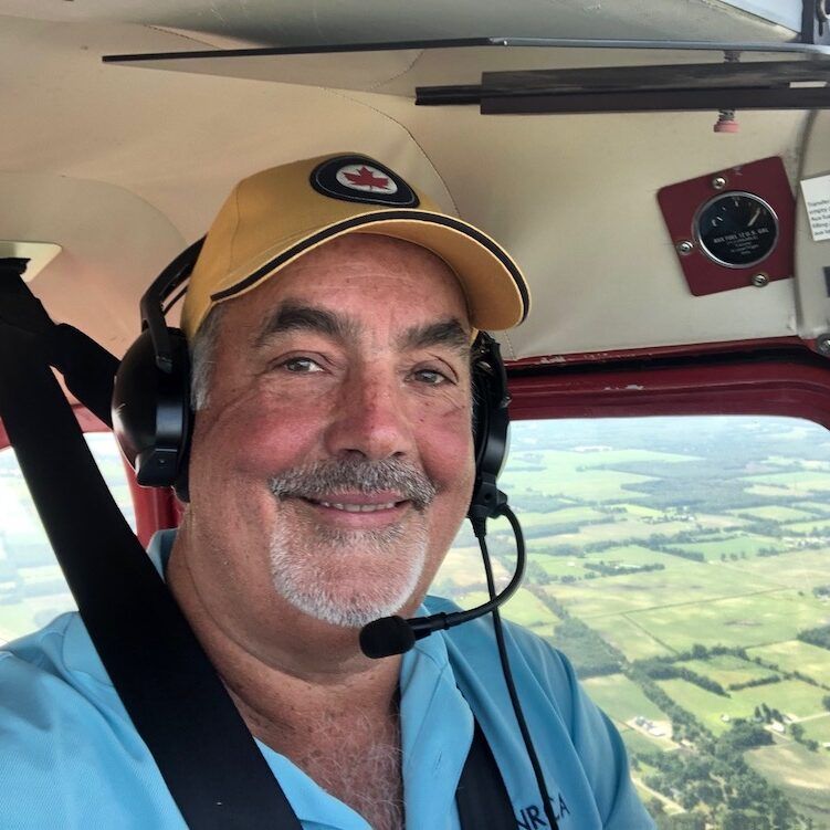 Man smiling in a small plane cockpit, wearing headset and hat, flying over green fields.