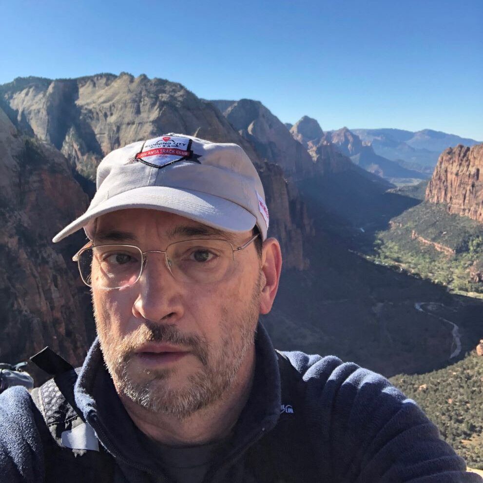 Man in hat and glasses with mountain vista in background.