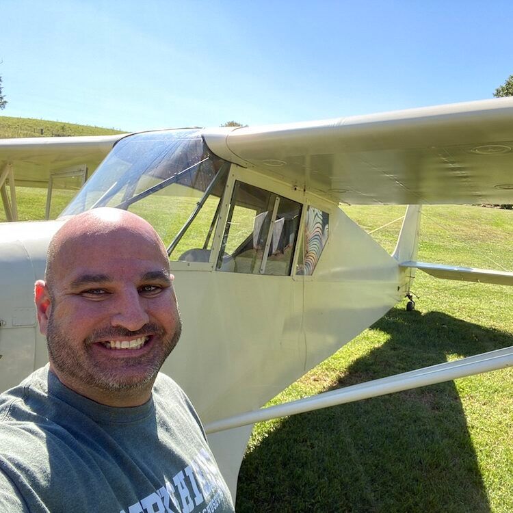 Man smiles in front of a light-colored small aircraft on a grassy field. Sunny, blue sky.