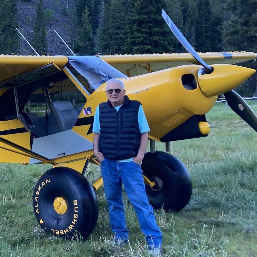 Man standing in front of yellow bush plane with large tires; wearing jeans, vest, and sunglasses.