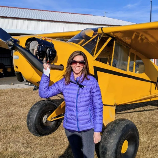 Man in jeans, plaid shirt, and vest, standing next to a blue and white airplane on a grassy airfield.