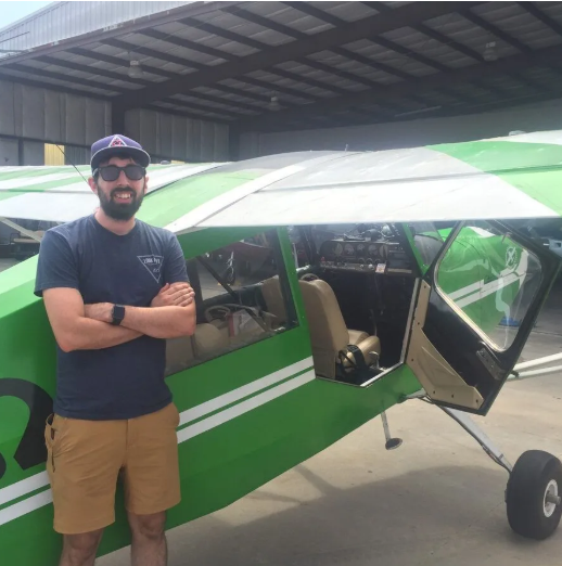 Man in front of brown and red airplane. Smiling with hands on hips. Airport setting.