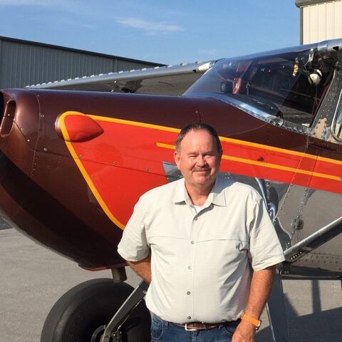 Man in front of brown and red airplane. Smiling with hands on hips. Airport setting.