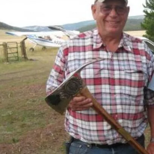 Man smiles in front of a light-colored small aircraft on a grassy field. Sunny, blue sky.