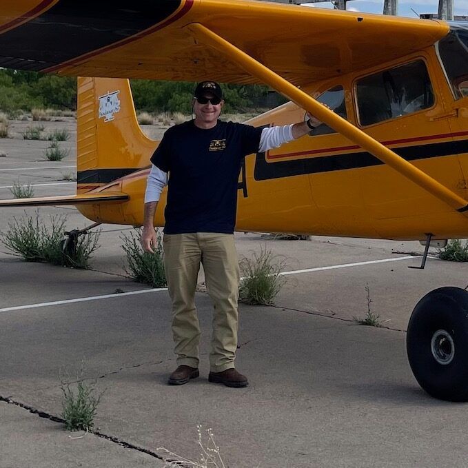 Man in dark blue shirt and khakis standing by a yellow airplane on a tarmac.