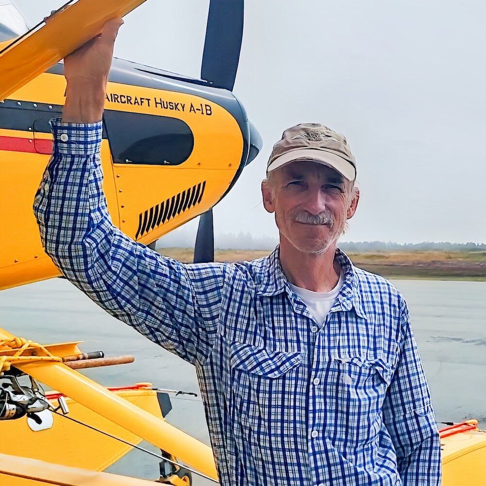 Man in plaid shirt and cap, next to a yellow floatplane, holding its wing.