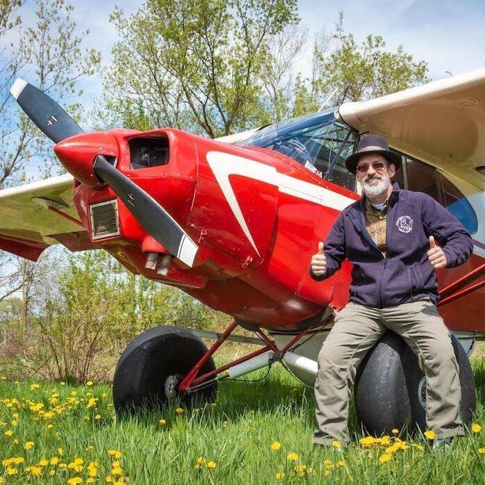 Man in hat sits on plane tire, giving thumbs-up in front of a red plane in a field.