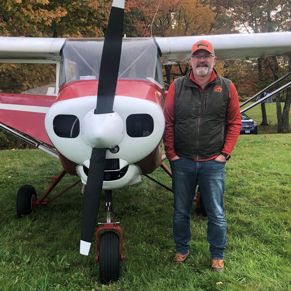 Man standing by a red and white airplane, smiling, wearing a vest, jeans, and a hat, outdoors in autumn.