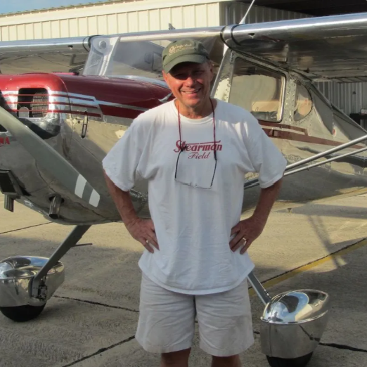 Man standing in front of yellow bush plane with large tires; wearing jeans, vest, and sunglasses.