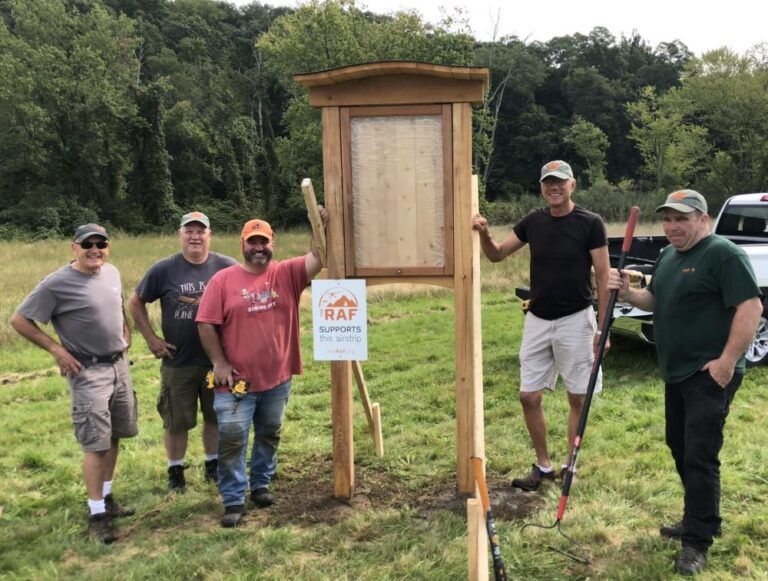 A group of men are standing next to a wooden sign in a field.