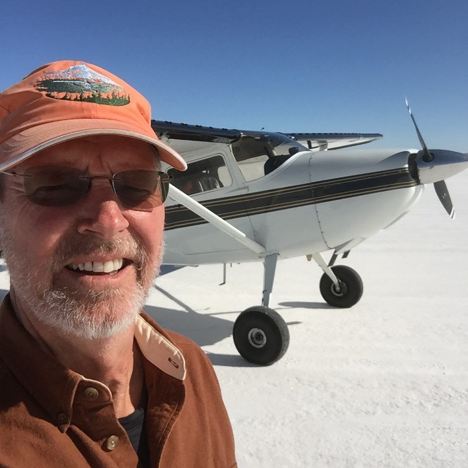 Man in hat and sunglasses in front of a small white airplane on a white plane. Blue sky.