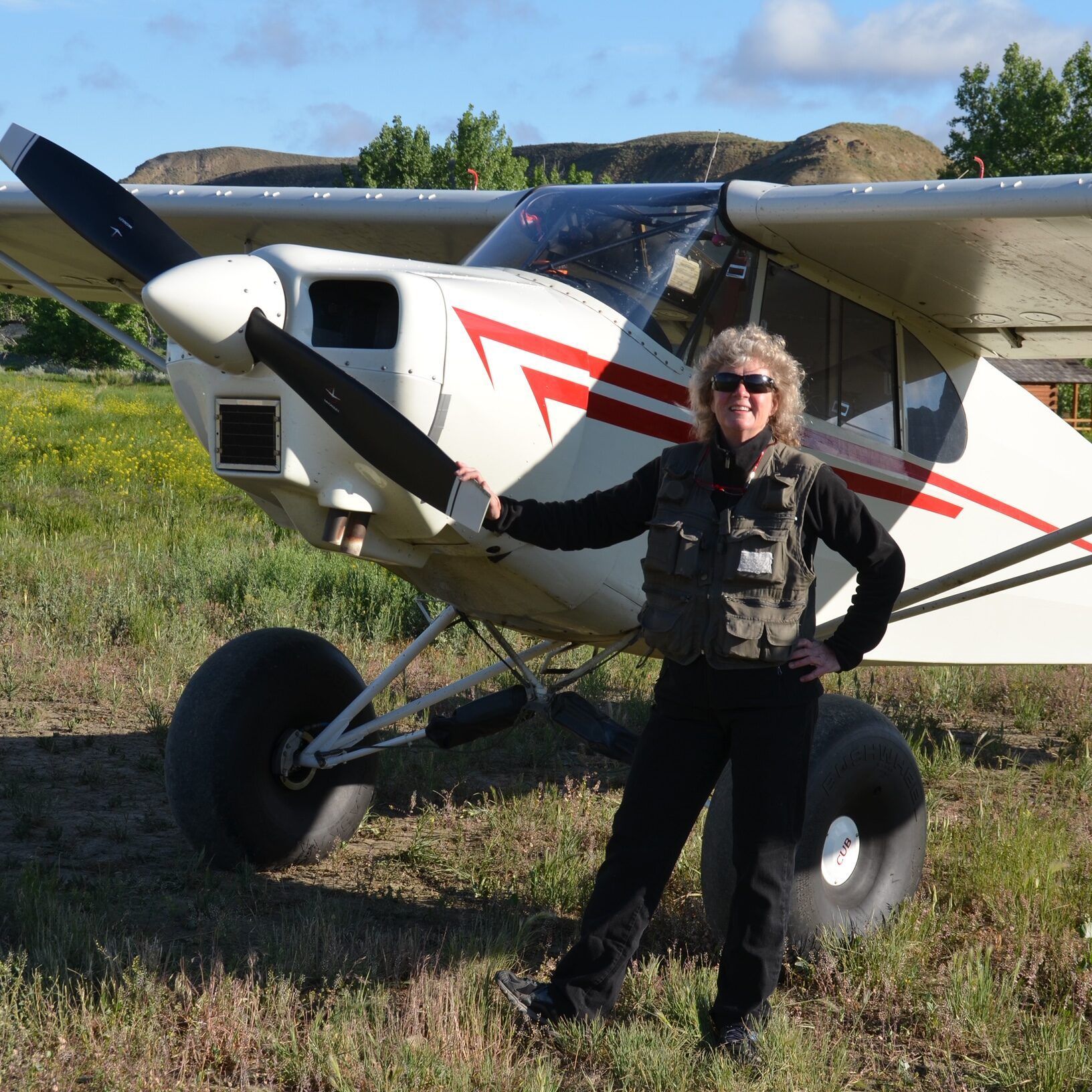 Woman in flight vest stands by a white and red airplane with oversized tires in a grassy field.