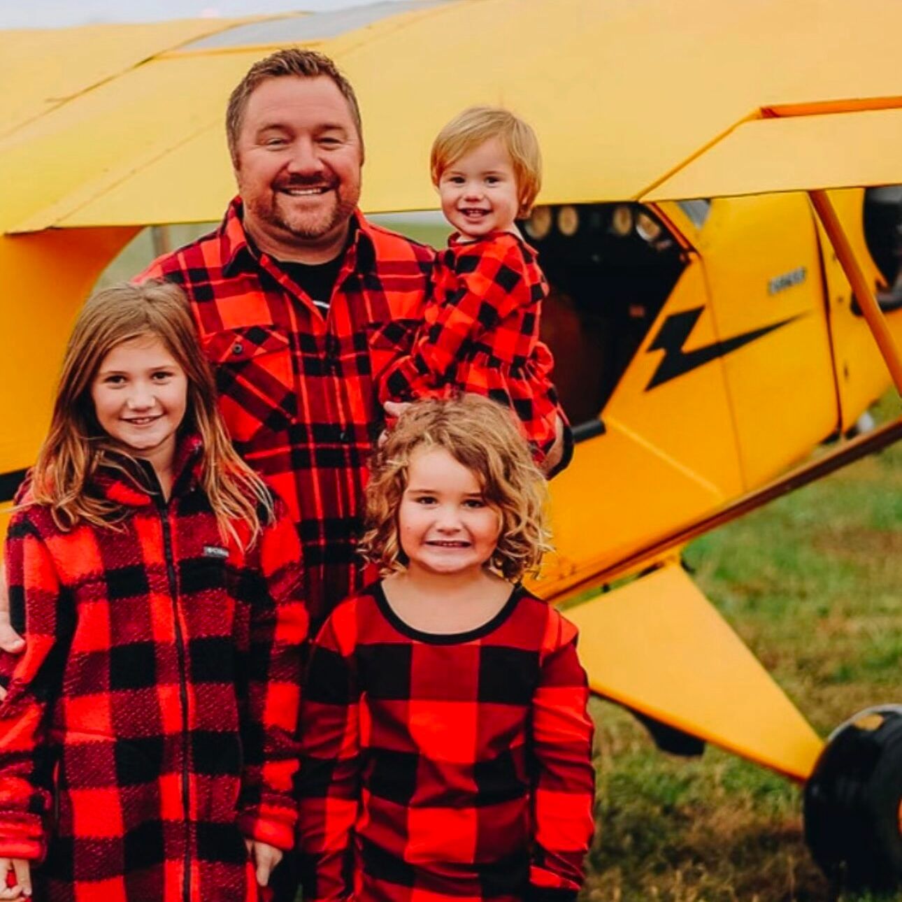Man and three young girls in matching red plaid pajamas pose by a yellow plane.