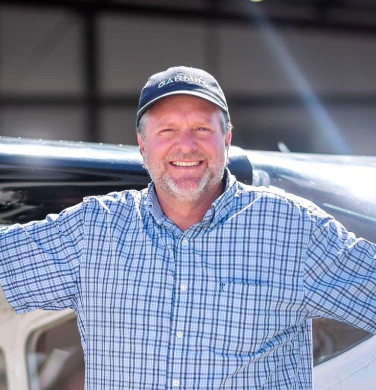 A man wearing a plaid shirt and a hat stands in front of an airplane