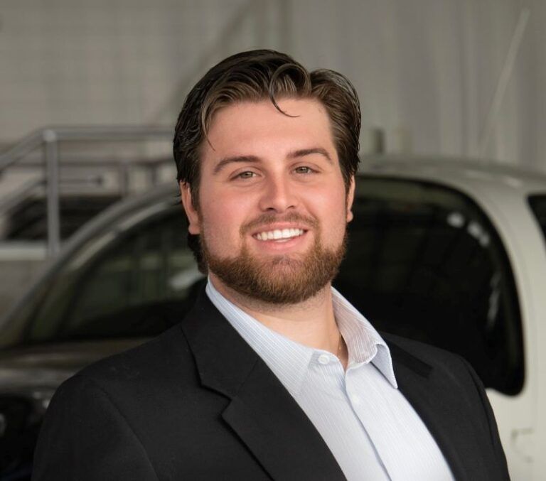 A man in a suit is smiling in front of a white car.