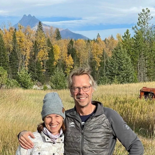 Man smiles in front of a light-colored small aircraft on a grassy field. Sunny, blue sky.