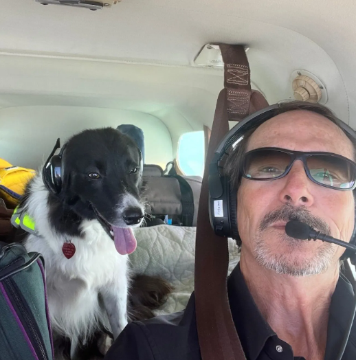 Man smiling in a small plane cockpit, wearing headset and hat, flying over green fields.