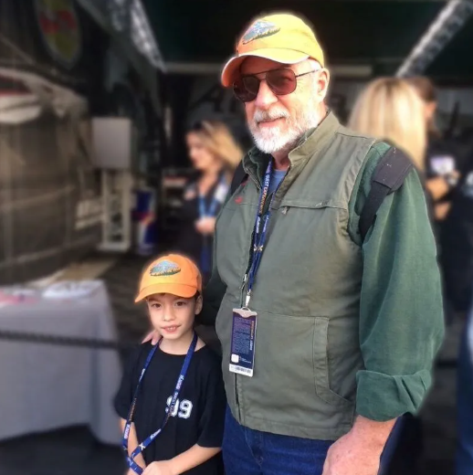 Man smiles in front of a light-colored small aircraft on a grassy field. Sunny, blue sky.
