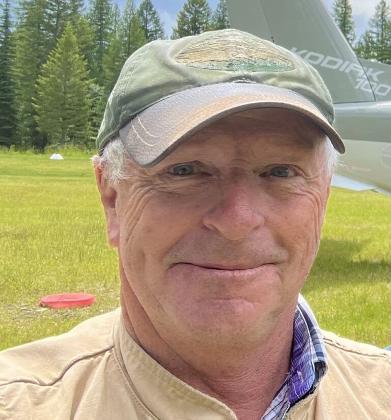 A man wearing a hat and a jacket is smiling in front of a plane.