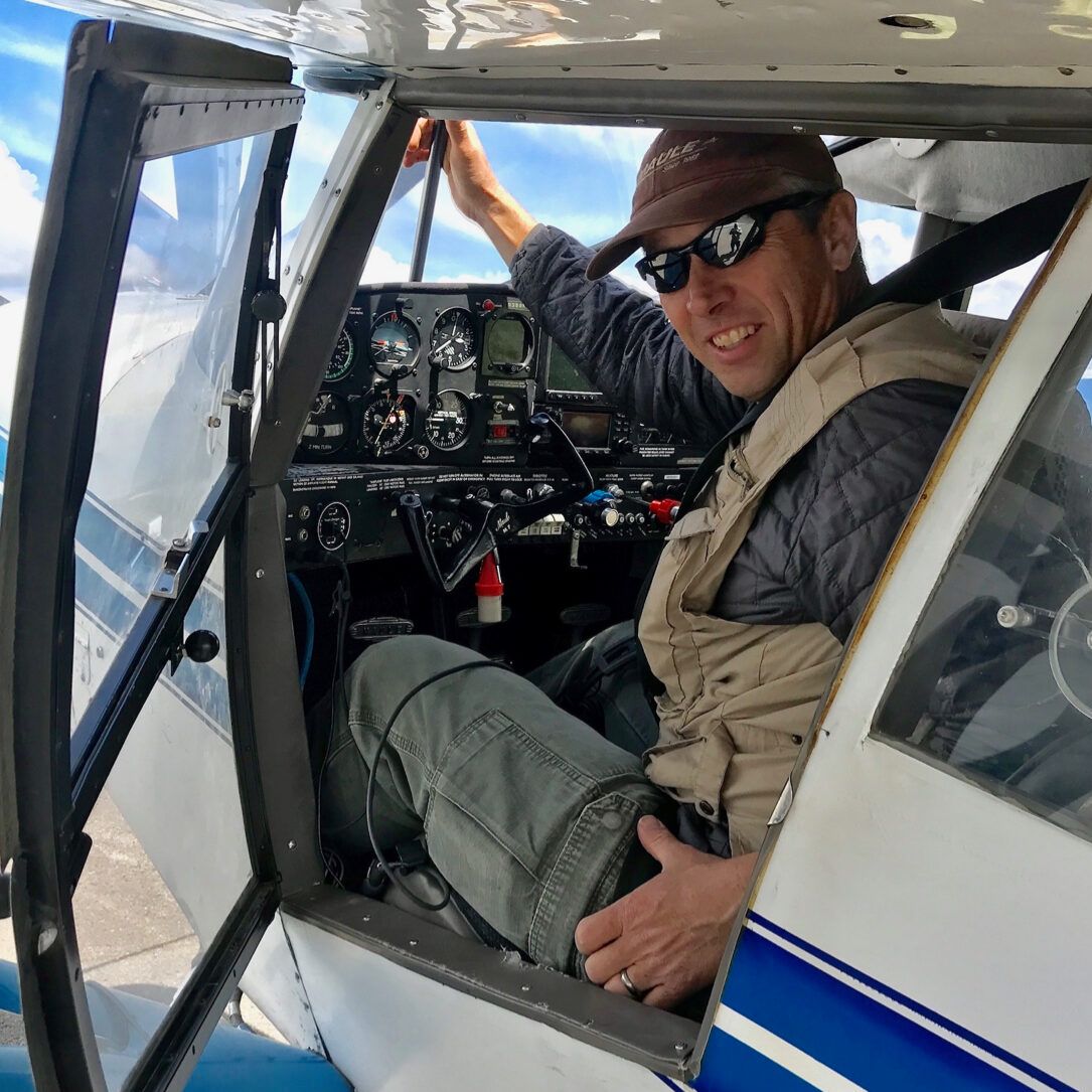 Man in sunglasses and hat seated in the cockpit of a small airplane, smiling.