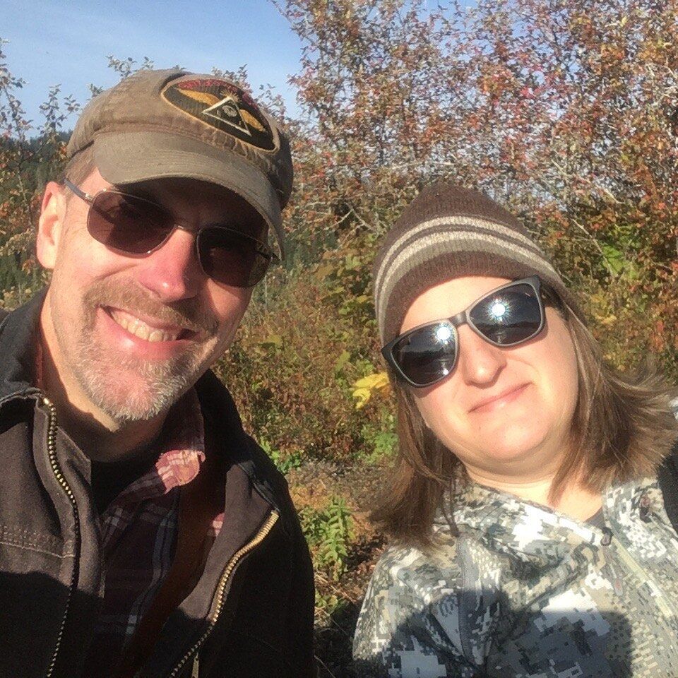 Man and woman in sunglasses smiling, in front of fall foliage. Man in cap, woman in striped beanie.