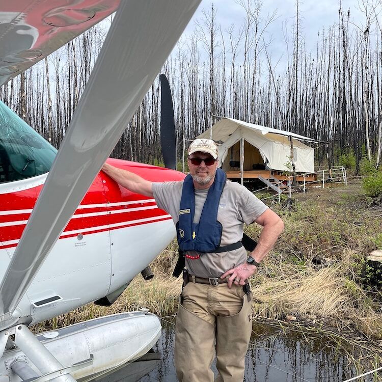 Man in life vest stands by a floatplane, next to a tent in a forest.