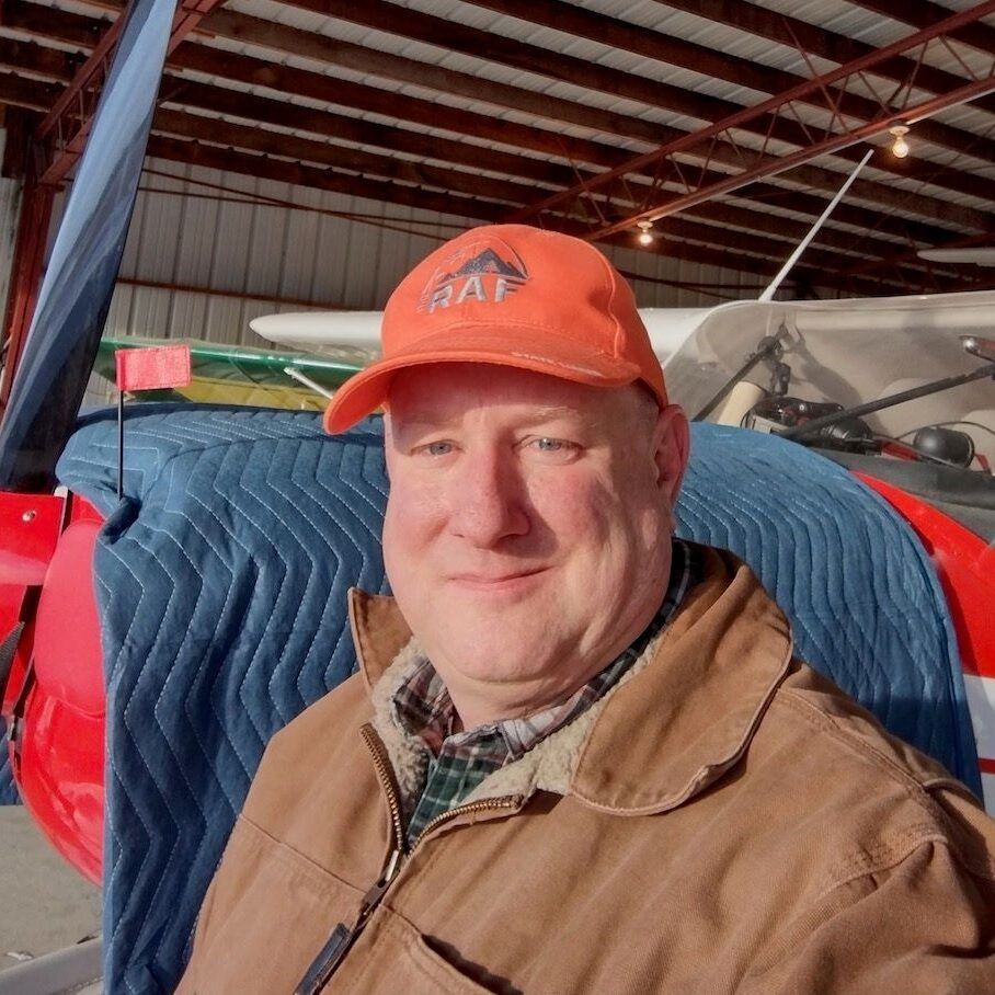 Man wearing orange cap and brown jacket, smiling in front of a small airplane in a hangar.