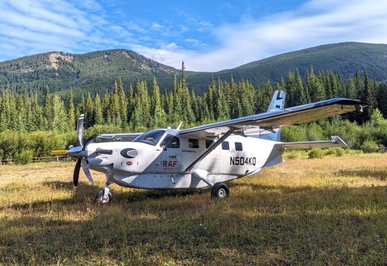 A small propeller plane is parked in a field with mountains in the background.