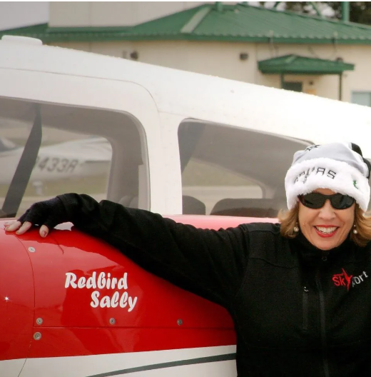 Man in black t-shirt smiles in front of a red and white airplane in a hangar.