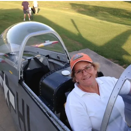 Man in jeans, plaid shirt, and vest, standing next to a blue and white airplane on a grassy airfield.